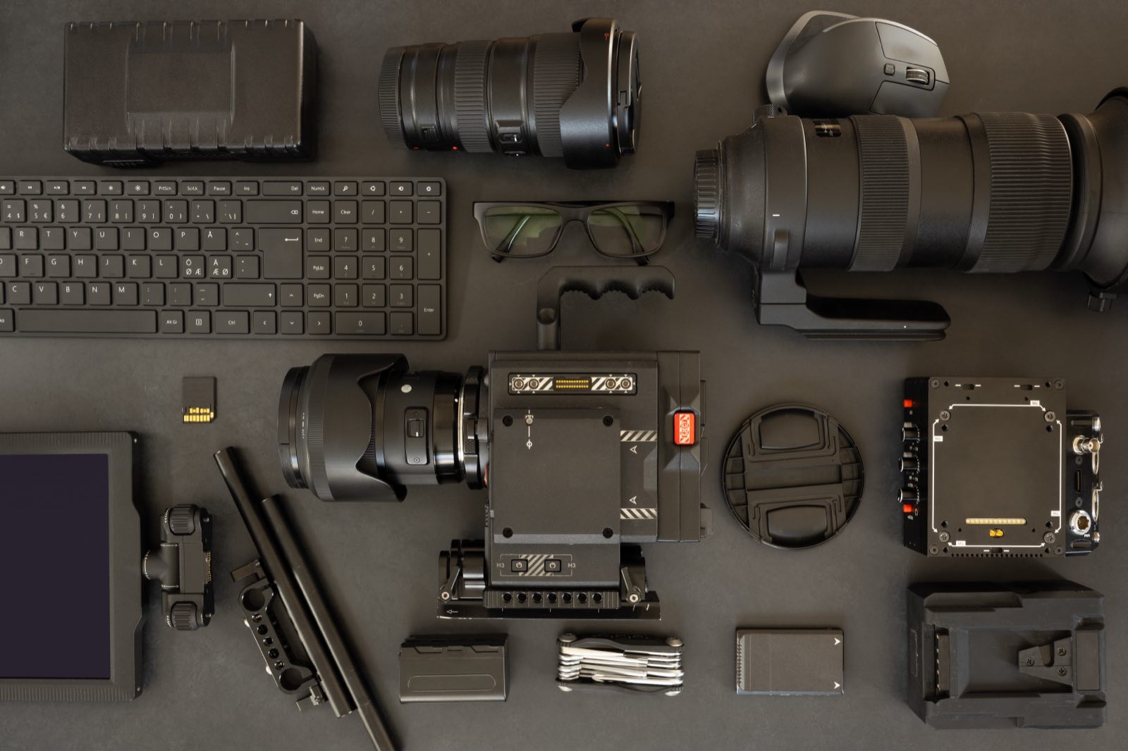 A videographer's kit laid out on a dark surface: a cinema camera on its rig, long lenses, a monitor, batteries and media cards